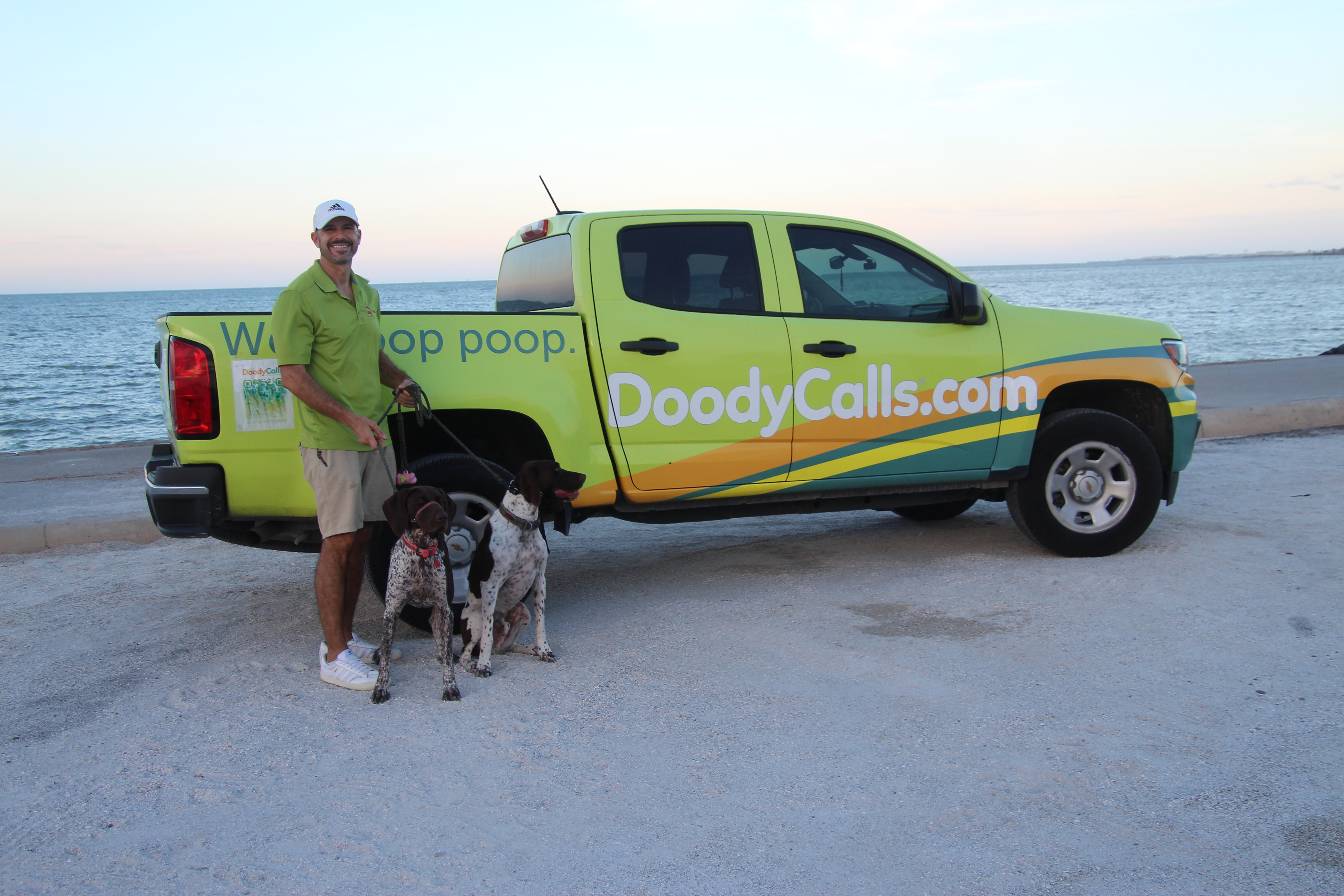 Photo of Chris and two dogs infront of a Doody Calls pick up truck on the beach